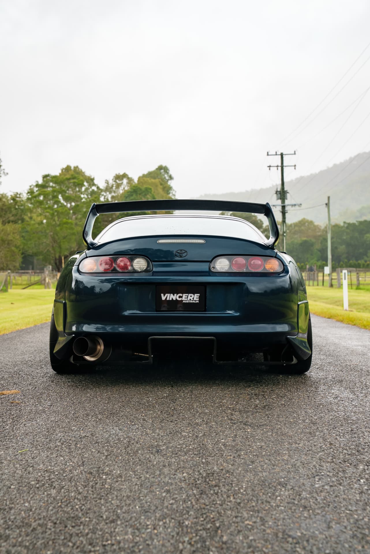 Rear of a teal Supra with large wing on a wet road